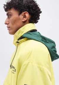 Νέος με σγουρά μαλλιά που φοράει κίτρινο μπουφάν με πράσινη κουκούλα, κοιτώντας πλάγια ενάντια σε ένα απλό φωτεινό φόντο.