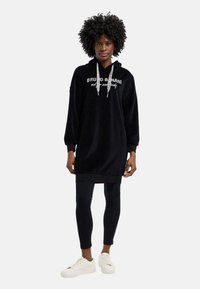 Schwarzer Kapuzenpullover aus weichem Material mit langen Ärmeln, ausgestattet mit einem Logo-Druck auf der Vorderseite und einem Kordelzug. Kombiniert mit schwarzen Leggings und weißen Sneakers.