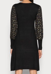 Robe noire à longues manches bouffantes ornée de pois métalliques dorés. Texture lisse, décolleté rond et silhouette en A.