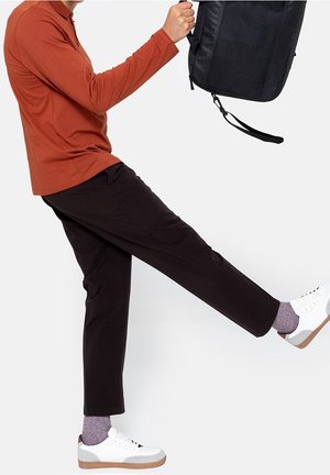 Persoon in een roestkleurig shirt met lange mouwen, zwarte broek, paarse sokken en witte sneakers tilt een been op terwijl hij een zwarte rugzak vasthoudt tegen een witte achtergrond.