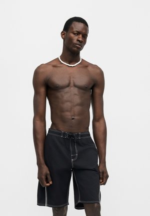 Homme torse nu portant un short de bain noir avec des coutures blanches et un collier de perles blanc, debout devant un fond blanc uni.