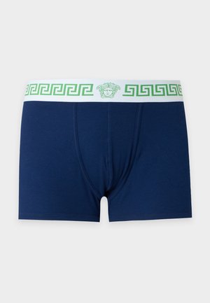 Hombre sin camiseta con cabello oscuro que lleva calzoncillos bóxer azul marino con una cintura blanca que presenta un patrón geométrico verde, de pie descalzo sobre un fondo blanco.
