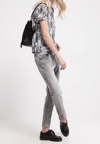 Top de manga corta en blanco y negro con patrón, jeans ajustados gris claro y zapatos negros con cordones. Mochila negra llevada sobre un hombro.