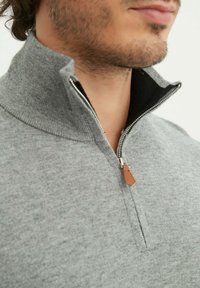 Gilet zippé gris à col haut, avec une doublure noire contrastante et un accent en cuir marron sur le tirette de la fermeture éclair. Texture douce.