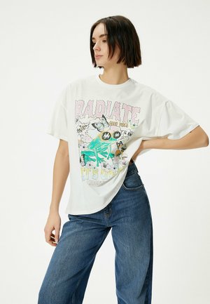 Hvid oversized t-shirt med et farverigt frø-grafik, sommerfugle og tekst der står "RADIATE GOOD VIBES" i pink og grøn.