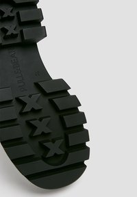 Semelle de bottes en caoutchouc noir présentant un motif de bande de roulement texturé avec des blocs en relief et des formes en "X", marquée du texte "PULL&BEAR" et de la taille 37.