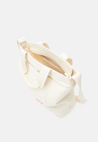 Torba tote wykonana z płótna w kremowym kolorze z dwoma górnymi uchwytami i beżowymi paskami. Posiada czerwone logo oraz otwarty górny z przestronnym wnętrzem.