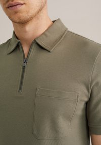 Polo de color verde oliva con cuello con cremallera, mangas cortas y un bolsillo en el pecho izquierdo con costuras limpias y textura suave.