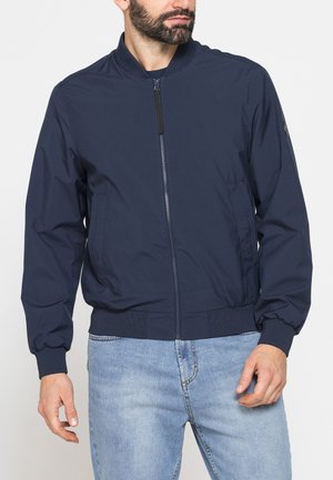 Uomo che indossa una giacca bomber blu navy con zip e jeans azzurri, in piedi con le mani rilassate lungo i fianchi.