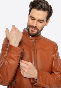 Giacca in pelle marrone con colletto alto, chiusura frontale con zip e tasche con zip. La texture è liscia con dettagli di cucitura visibili.