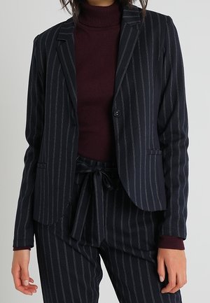 Blazer rayé en bleu marine avec des lignes blanches, coupe ajustée, fermeture à un bouton et poches latérales ; associé à un col roulé bordeaux.