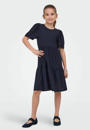 Ragazza giovane in piedi con una mano sul fianco, indossa un vestito midi nero a trafori e scarpe Mary Jane nere, sorridendo verso la fotocamera su sfondo semplice.