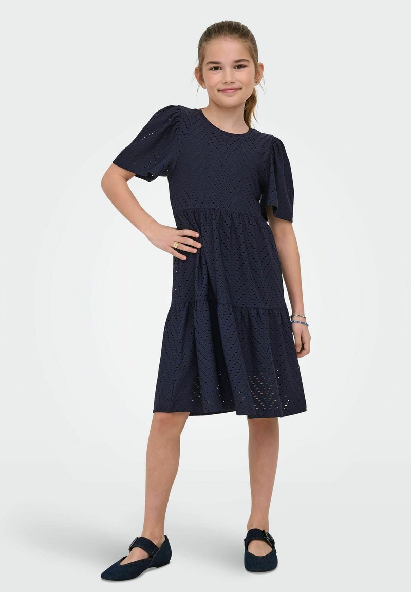 Młoda dziewczyna stoi z jedną ręką na biodrze, ubrana w czarną sukienkę midi z ażurowymi wzorami i czarne czółenka Mary Jane, uśmiecha się do aparatu na jednolitym tle.