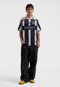 Polo majica s kratkim rukavima u crno-bijelim prugama s plavim logotipom i ovratnikom; uparena s širokim crnim trapericama i žutim tenisicama.