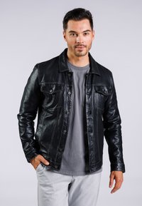Chaqueta de cuero negra con diseño de botones, que presenta cuatro bolsillos frontales y costura visible. Llevada sobre una camiseta gris.