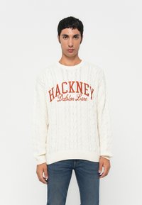 Krem pulover pleten s kabli, ki v oranžni barvi nosi napis "HACKNEY Dalston Lane". S prožnimi manšetami in robom; sproščen kroj. Nosi se z modrimi džinsom.