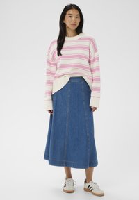 Femme portant un pull rayé blanc et rose, une jupe midi en denim bleu, et des baskets blanches à rayures noires, posant devant un fond uni.