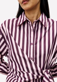 Frau trägt ein pinkes und lila gestreiftes, hochgeschlossenes Hemd, das an der Taille zu einem Knoten gebunden ist, abgebildet von Kinn bis Hüfte.