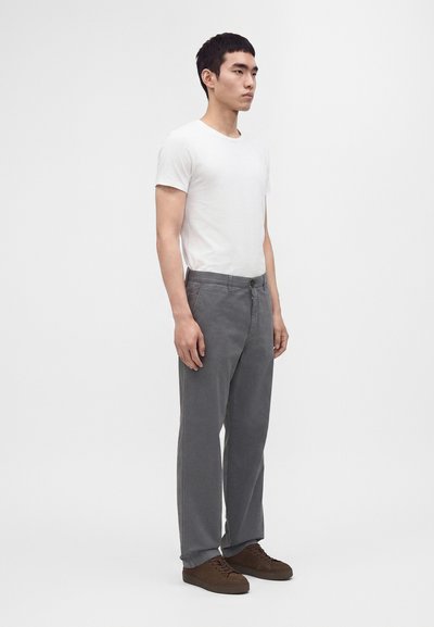 Jeune homme debout, tourné vers la droite, portant un t-shirt blanc à manches courtes, un pantalon gris et des chaussures marron sur un fond clair uni.
