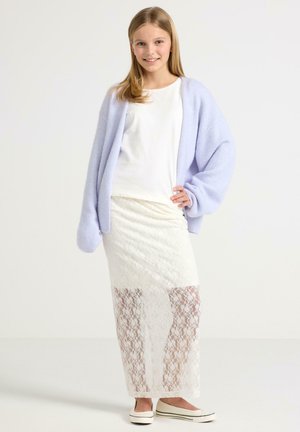 Nuori tyttö hymyilee, yllä valkoinen toppi, liilanvärinen ylisuurikokoinen neuletakki, pitkä valkoinen pitsihame ja valkoiset liukusandaalit, seisoo yksivärisellä taustalla.