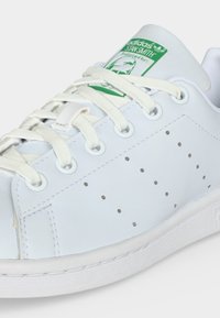 Biele tenisky Adidas Stan Smith s bielymi šnúrkami, perforovanými bočnými dierami a zeleným logom na jazyku.