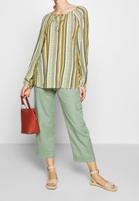 Blouse à manches longues rayée en jaune et vert avec un décolleté en goutte. Associée à un pantalon cargo vert et un sac à main rouge ; sandales beiges.
