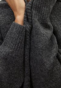 Handen die de kraag van een donkergrijze, geribbelde breisweater aanpassen, waarbij de zachte textuur en het gezellige stof worden benadrukt.