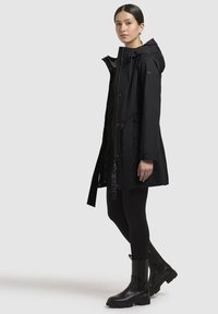 Schwarze wasserdichte Parka mit Kapuze, Reißverschluss und Druckknöpfen; mit strukturierten Einsätzen und Seitentaschen; kombiniert mit schwarzen Leggings und Stiefeln.