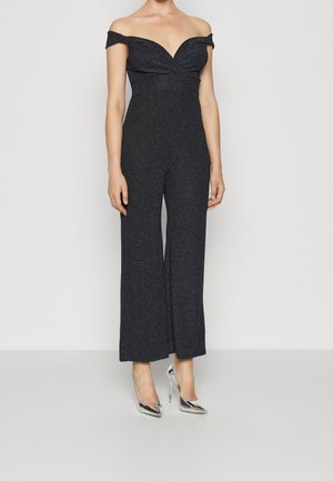 Zwarte jumpsuit zonder schouders, met een aansluitende lijfje, wijde pijpen en een subtiele glans. Gecombineerd met zilveren hoge hakken.