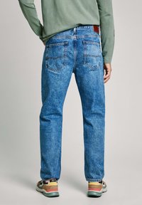 Vaqueros de denim azul con un diseño de pierna recta, con bolsillos traseros cosidos y un parche de cuero. Combinados con zapatillas de colores y una camisa verde.