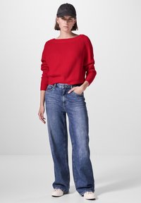 Roter gerippter Pullover, lockere Passform, kombiniert mit hellblauen weiten Jeans. Das Modell trägt eine dunkle Mütze und weiße Sneaker. Einfaches, lässiges Ensemble.