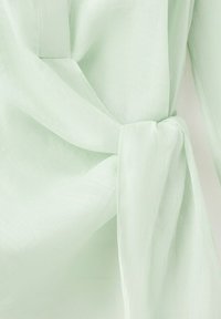 Tela verde claro atada en un nudo suelto, destacando su textura suave y sutiles detalles de costura en una prenda.