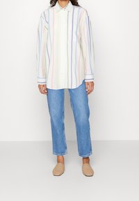 Chemise à rayures dans des couleurs pastel (rose, bleu, jaune) avec une poche poitrine, associée à un jean droit bleu et des chaussures slip-on beiges.