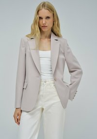 Blazer beige chiaro con un design sartoriale, colletto appuntito, chiusura a un bottone e una sottile trama a righe, abbinato a pantaloni bianchi.