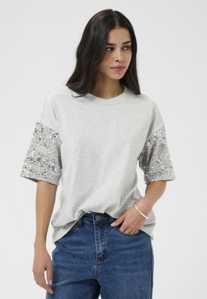 Hosszú sötét hajú nő, aki világosszürke pólót visel ezüst flitteres ujjal és kék farmert, egyik kezét zsebében tartva áll.