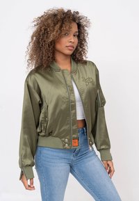 Veste bomber en satin vert olive avec fermeture éclair à l'avant, poignets côtelés et détails brodés. Associée à un jean en denim bleu clair.