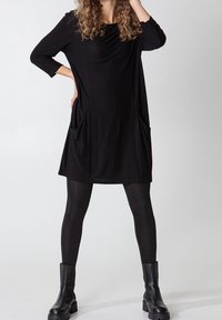 Svart knälång klänning med trekvartsärmar och stora sidofickor, tillverkad av mjukt tyg, kombinerad med svarta leggings och kraftiga stövlar.