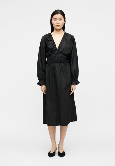 Γυναίκα όρθια, φοράει μαύρο μίντι φόρεμα με μακριά μανίκια, βαθύ V-λαιμόκοψη και πιέτες στη μέση, συνδυασμένο με μαύρα μυτερά παπούτσια, σε απλό λευκό φόντο.