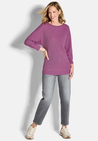 Lila gerippter Strickpullover mit weitem, lockeren Ausschnitt und langen Ärmeln, kombiniert mit hellgrauen, geraden Jeans und beigen Sneakers.