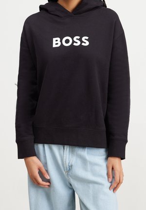 Person trägt einen schwarzen Kapuzenpullover mit dem weißen Schriftzug BOSS auf der Brust, kombiniert mit hellblauen Jeans.