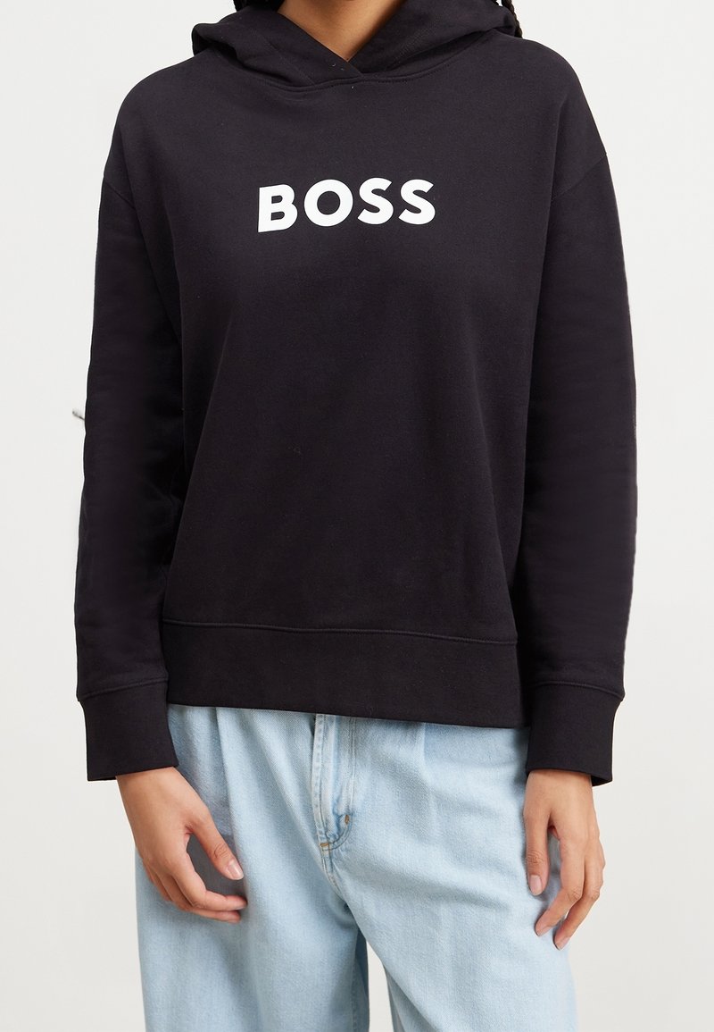 Personne portant un sweat à capuche noir avec le mot BOSS en blanc sur la poitrine, associé à un jean bleu clair.