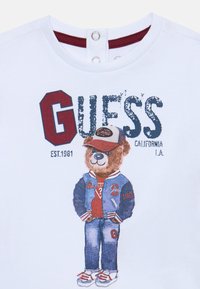 Hvid t-shirt med et grafisk motiv af en bjørn iført en blå varsityjakke, rød skjorte og jeans. Har "GUESS" branding i marineblå og vinrød.