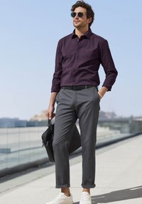 Purple button-up shirt, gray patterned trousers, white sneakers, and black bag, standing against a clear sky and urban background.