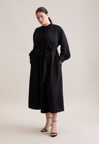 Schwarzes Kleid mit hohem Kragen, langen Ärmeln, elastischem Gathering, Taillengürtel und seitlichen Taschen; kombiniert mit schwarzen, verzierten High Heels.