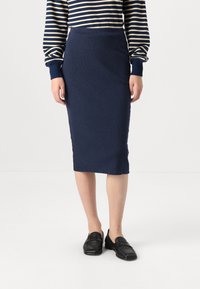 Jupe crayon bleu marine en tissu tricoté côtelé, avec une silhouette ajustée, s'étendant jusqu'au mollet. Associée à des mocassins noirs.