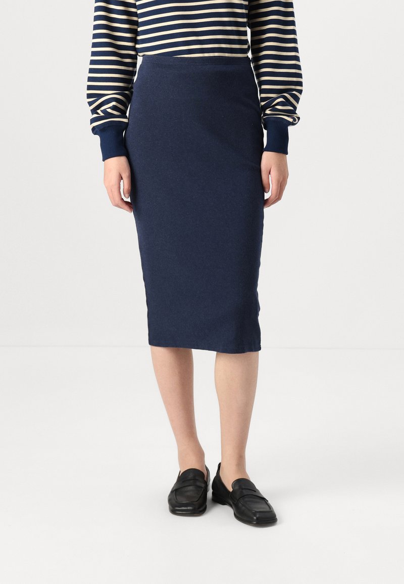 Jupe crayon bleu marine en tissu tricoté côtelé, avec une silhouette ajustée, s'étendant jusqu'au mollet. Associée à des mocassins noirs.