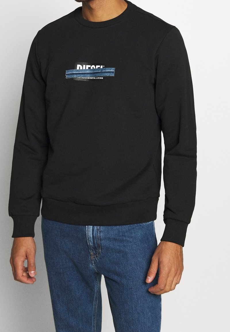 Homme portant un sweat-shirt Diesel noir avec un jean bleu, logo Diesel partiellement recouvert par une bande bleue sur la poitrine.