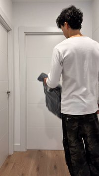 En person som bär en vit långärmad tröja och svarta kamouflagebyxor håller ett vikt grått plagg och står i en modern korridor med vita dörrar.