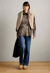 Chemise à motifs animaliers, gilet beige, jeans bleus et chaussures à plateforme beiges. Tenant un sac noir. Arrière-plan simple avec un sol en bois.