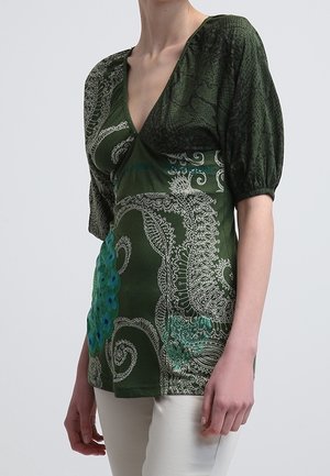 Vrouw draagt een groene, met patroon bedrukte wikkelblouse met geplooide korte mouwen en witte broek, staand tegen een effen achtergrond.